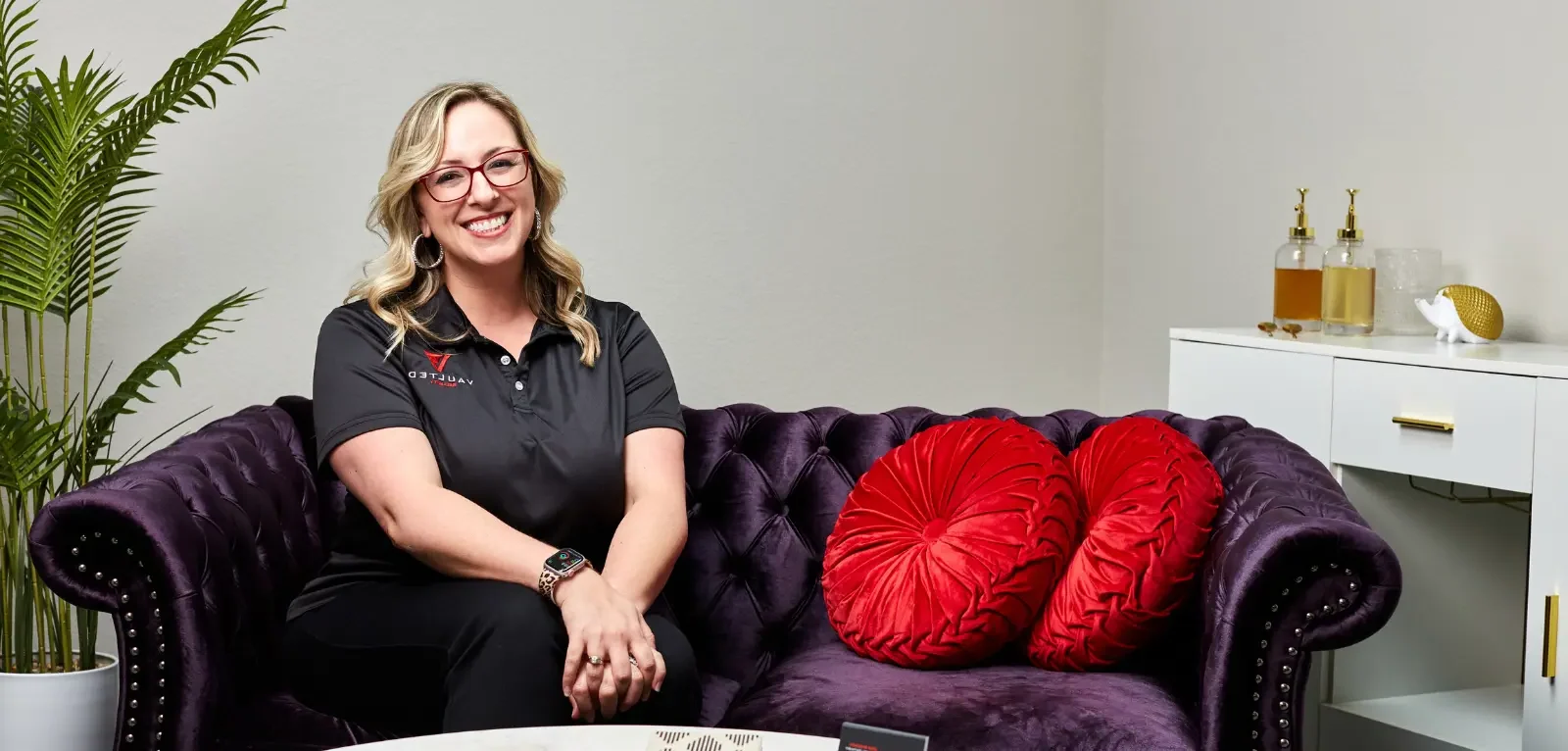 Woman in Vaulted Security polo sits on black couch with red pillows.