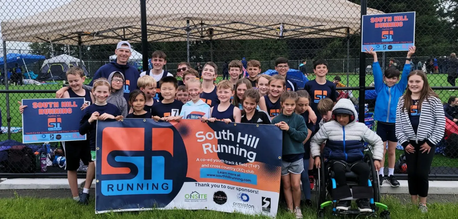 A group of children in South Hill Running Club t-shirts hold up a banner and posters to cheer on South Hill Running Club.